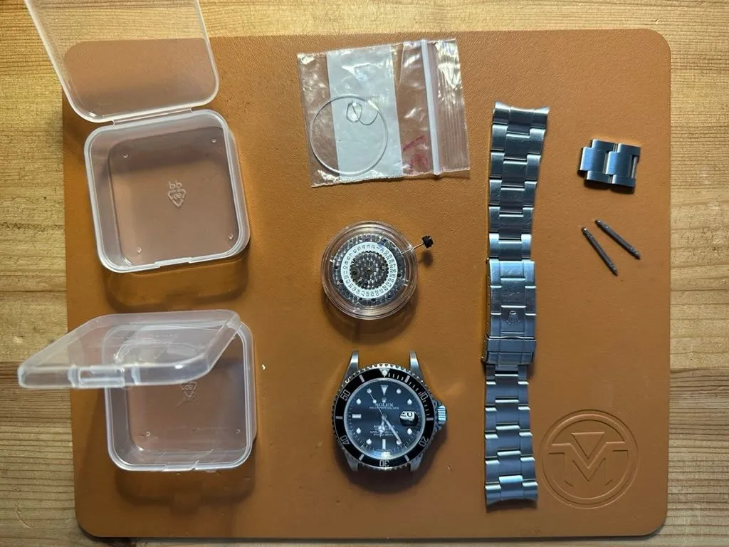 Parts for the 16610 build laid out on a leather mat: watch head, spare movement, bezel parts, crystal, bracelet and spring bars